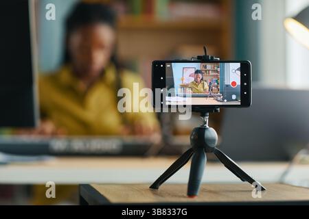 Frau, die ein Smartphone auf einem Stativ für die Aufnahme von Videoinhalten im Home Office verwendet. Hintergrund verschwommen, wodurch der Fokus auf das Gerät für Profis gelegt wird Stockfoto