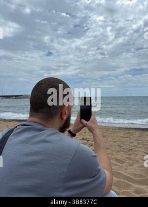 Der weiße Typ, Tourist, macht Fotos vom Meer auf seinem Telefon, das am Ufer sitzt, vertikales Foto Stockfoto