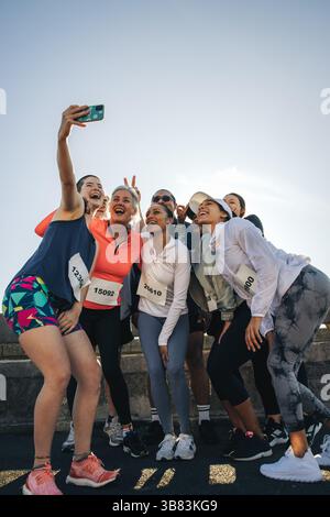 Eine Gruppe von Freunden aus einem Laufclub trifft sich zu einem fröhlichen Selfie während eines Outdoor-Events. In Sportkleidung und Wettkampflätzchen bekleidet, strahlen sie happ aus Stockfoto
