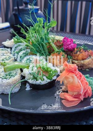 Lebendige Sushi-Platte mit frischem Gemüse, Reisbrötchen und Algen auf einem dekorativen grauen Teller mit Tee-Set und Holzelementen. Stockfoto