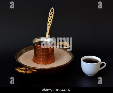 Kupfertürke in einem Tablett mit Sand und einer Tasse frisch gebrühten schwarzen Kaffee auf schwarzem Hintergrund. Nahaufnahme. Stockfoto