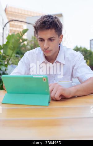 Junger Mann, der ein Tablet an einem Café-Tisch im Freien in vertikaler, urbaner Umgebung benutzt Stockfoto