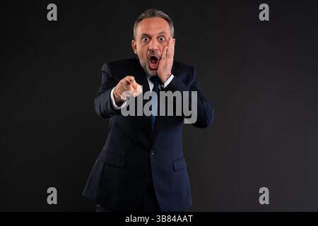 Überraschter Geschäftsmann im Anzug, der mit Hand auf Gesicht direkt auf die Kamera zeigt und Schock, Erkenntnis oder Schuld ausdrückt. Studio auf schwarzem Hintergrund. Stockfoto