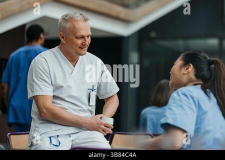 Reifer männlicher Arzt, der mit einem Kollegen spricht, während er in der Krankenhauskantine auf dem Tisch sitzt Stockfoto