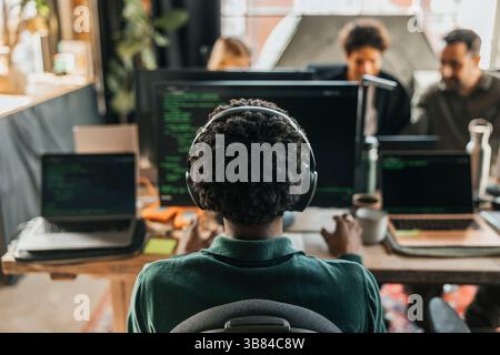 Rückansicht eines männlichen Computerprogrammierers mit lockigen Haaren, der auf dem Desktop-PC kabellose Kopfhörercodierung trägt, während er auf dem Cha sitzt Stockfoto