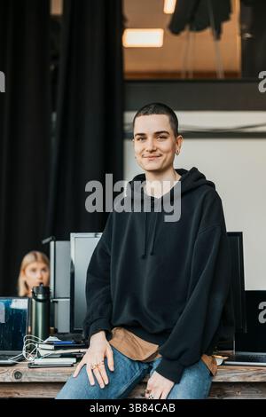 Porträt eines lächelnden, nicht binären IT-Profis, der am Arbeitsplatz auf dem Schreibtisch sitzt Stockfoto