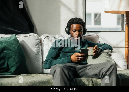 Junger Geschäftsmann, der Musik über kabellose Kopfhörer hört, während er sein Smartphone im Büro auf dem Sofa sitzt Stockfoto