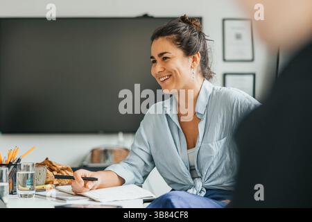Glückliche junge Geschäftsfrau, die im Tagebuch schreibt, während sie mit einem Kollegen im Amt spricht Stockfoto