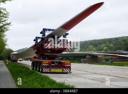 Der rot-weiße Lkw fährt mit einem sehr langen Windturbinenschaufel auf einem speziellen Anhänger auf einer Landstraße neben einem Lagerbereich mit anderen Stockfoto