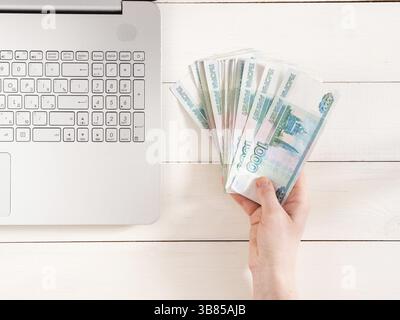 Hand hält russische Rubel in der Nähe von Laptop auf weißem hölzernem Hintergrund. Draufsicht oder flache Lagen. Finanz- und Ertragskonzept Stockfoto