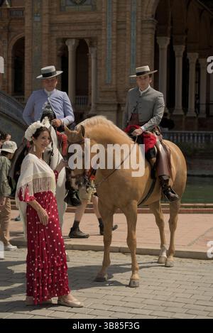 Andalusische Fahrer und eine Frau in rotem Flamenco-Kleid auf der Plaza de España während der Feria de Sevilla 2025 in Sevilla, Andalucía, Spanien. Stockfoto