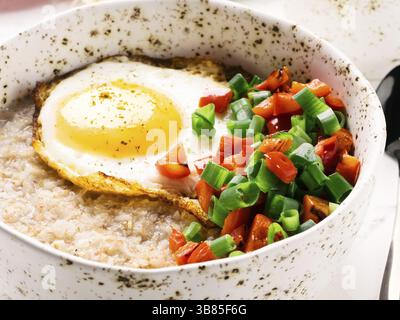 Herzhaftes Haferbrei in handgefertigter trendiger Schüssel, serviert mit Gemüse und Spiegelei auf weißem Betonhintergrund. Rote Paprika und Zwiebeln auf dem Hintergrund Stockfoto