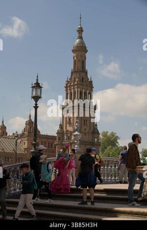 Menschen in traditionellen Flamenco-Kleidern und Touristen, die an der Plaza de España während der Feria de Sevilla 2025, Sevilla, Andalucía, Spanien vorbeilaufen. Stockfoto