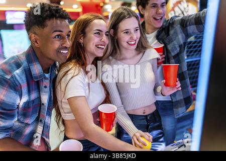 Eine Gruppe fröhlicher, junger, multiethnischer Freunde, die Spaß miteinander haben, Spielautomaten spielen und rote Tassen in einer Bowlingbahn halten Stockfoto
