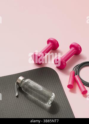Stylisches Fitness-Training und Fitnesskonzept in Grau und Pink. Perspektivische Ansicht von grauer Sportmatte, Wasserflasche, rosa Hanteln und Springseil auf rosa Bac Stockfoto