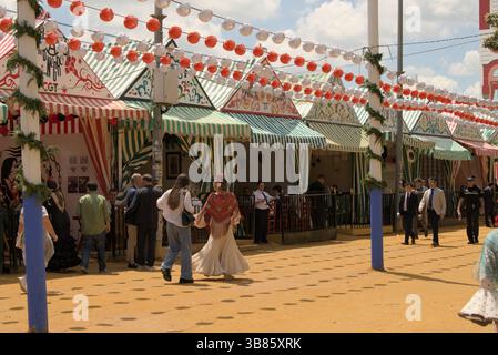 Besucher, die während der Feria de Sevilla 2025, Sevilla, Andalucía, Spanien, an bunten gestreiften Kasetten und Laternen vorbeispazieren. Stockfoto