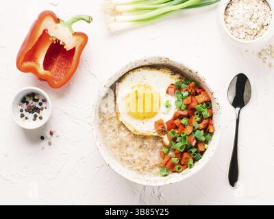 Schmackhafte Haferflocken in Handwerk trendige Schüssel, serviert mit Gemüse und Spiegelei auf weißem Beton Hintergrund. Rote Paprikaschote und Haufen Zwiebeln auf Hintergrund. Stockfoto