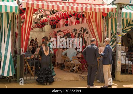 Elegante Besucher, die während der Feria de Sevilla 2025, Sevilla, Andalucía, Spanien, Gespräche vor einer gestreiften Caseta genießen. Stockfoto
