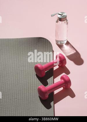 Stylisches Fitness- und Fitnesskonzept in Grau und Pink. Perspektivische Ansicht der grauen Sportmatte, der Wasserflasche und der pinkfarbenen Kurzhanteln auf pinkfarbenem Hintergrund. Einstellen für Stockfoto