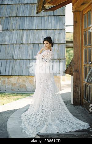 Braut in einem langen weißen Korbkleid steht an der Schwelle eines Holzhauses. Stockfoto