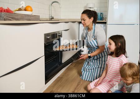 Eine Mutter nimmt frisch gebackene Croissants aus dem Ofen, während ihre Kinder eifrig zusehen. Stockfoto