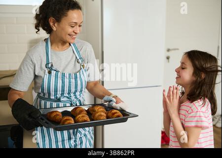 Eine lächelnde Mutter serviert frisch gebackene Croissants in einer gemütlichen Küche ihrer entzückten Tochter. Ein Moment familiärer Liebe und hausgemachter Kulinarik Stockfoto