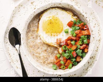 Schmackhafte Haferflocken in Handwerk trendige Schüssel, serviert mit Gemüse und Spiegelei auf weißem Beton Hintergrund. Rote Paprikaschote und Haufen Zwiebeln auf Hintergrund. Stockfoto
