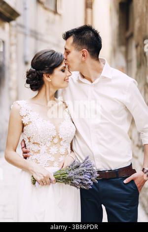 Sibenik, Kroatien - 05.06.17: Bräutigam umarmt und küsst Braut in einer Stirn in einem weißen Spitzenkleid mit einem Strauß Lavendel in ihren Händen auf der Straße von Stockfoto
