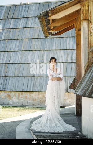 Braut in einem Korbkleid steht an der Schwelle eines Holzhauses. Stockfoto