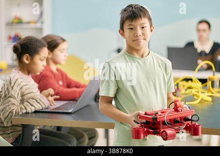 Porträt eines Jungen, der ein Robotermodell hält, in einer Klassenzimmer-Umgebung steht und fokussierte Schüler im Hintergrund an Laptops arbeiten Stockfoto