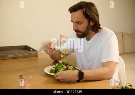 Ein Mann sitzt an einem Holztisch und genießt einen frischen grünen Salat in einer hellen, gemütlichen Atmosphäre. Die Umgebung ist einfach und doch elegant und betont gesunde Ernährung Stockfoto