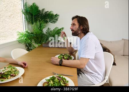 Ein Mann sitzt an einem Holztisch neben einem Freund und genießt eine frische Salatmahlzeit in einer hellen, heimeligen Umgebung mit natürlichem Licht und dekorativem Grün. Stockfoto