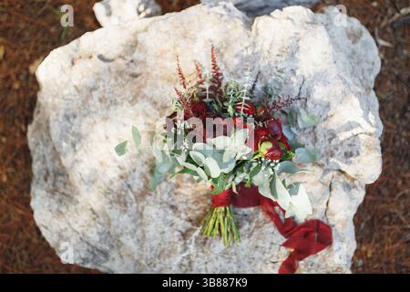 Brautstrauß aus weinroten Pfingstrosen, roten Rosen, Eukalyptuszweigen, eryngium, Baby Atem, Celosie und rote Bänder auf dem Stein. Stockfoto