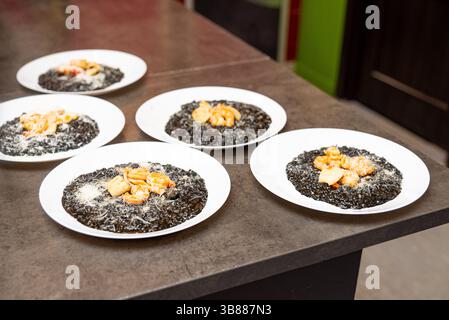 Mehrere Teller mit schwarzem Tintenfisch-Risotto, garniert mit gegrillten Garnelen und Jakobsmuscheln, zubereitet in einer professionellen Küche. Stockfoto