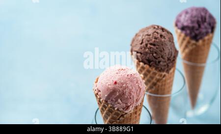 Verschiedene Eisbecher in Zapfen mit Kopierraum für Text oder Design. Bunte Eis in Zapfen Erdbeere, Schokolade, Heidelbeere auf blauem Hintergrund. Stockfoto