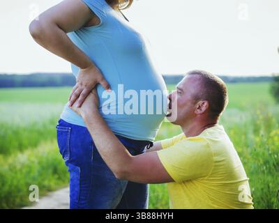 Ehemann küsst den Bauch seiner schwangeren Frau auf dem Feld. Zwangloses schwangeres Paar draußen, Bauch und Mannkopf aus der Nähe Stockfoto