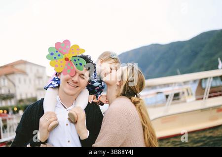 Mom küsst ihre Tochter. Der lächelnde Vater trägt seine Tochter auf den Schultern. Die Spielzeugblume bedeckte Daddy fast das Gesicht. Stockfoto