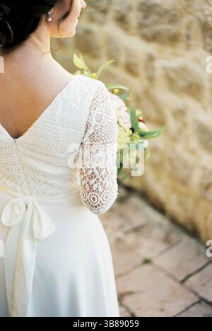 Braut in einem Spitzenkleid mit einem Blumenstrauß im Innenhof des Hauses. Rückansicht. Abgeschnitten. Stockfoto
