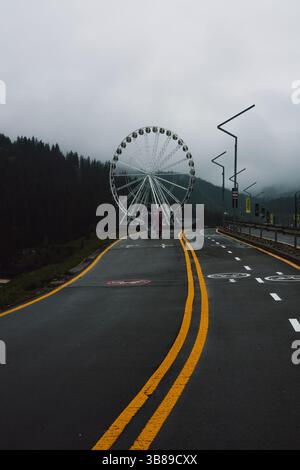Bukovel, Ukraine. 6. Mai 2025 zwei Figuren laufen auf einer Asphaltstraße auf ein Riesenrad zu, über dem bewölkter Himmel schwebt. Ein Straßenweg unter bewölktem Himmel. Stockfoto
