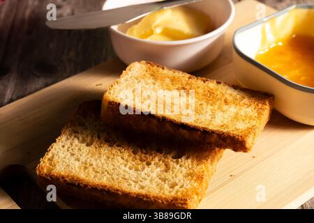 Geröstete Brotscheiben auf hölzernem Schneidebrett mit Butter und Marmelade, fertig zum Frühstück Stockfoto