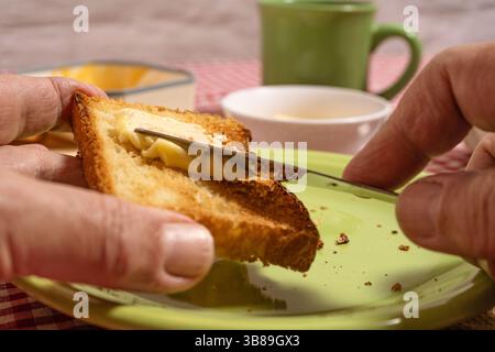 Nahaufnahme der Hände, die während des Frühstücks Butter auf Toast verteilen, mit Kaffeetasse und Butterschale im Hintergrund Stockfoto