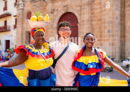Kolumbien, Cartagena - 16. April 2025: Junger Latino-Mann umarmt zwei fröhliche Afro-Latina Palenquera Frauen in bunten Kleidern und Obstkörben in Cartagena Stockfoto