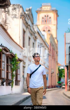 Latino-Mann mit weißem T-Shirt und beigefarbener Hose geht selbstbewusst und lächelnd durch farbenfrohe Kolonialstraßen in Cartagena, Kolumbien. Stockfoto