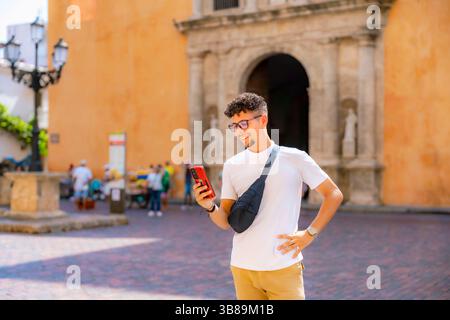 Junger Latino-Mann in weißem Hemd und khakifarbener Hose steht lächelnd mit Telefon in der Hand auf einem sonnigen plaza in Cartagena, Kolumbien. Stockfoto