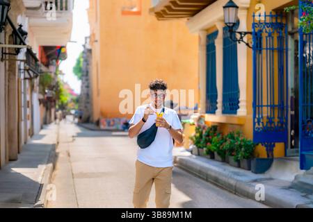 Latino-Mann mit weißem Hemd und khakifarbenen Hosen isst Mango freudig, während er eine farbenfrohe Straße in Cartagena, Kolumbien, hinunterschlendert. Stockfoto
