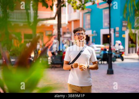 Latino-Mann mit weißem Hemd und khakifarbenen Hosen isst Mango freudig, während er eine farbenfrohe Straße in Cartagena, Kolumbien, hinunterschlendert. Stockfoto