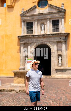 Ein glücklicher und entspannter junger Latino-Mann mit grauem Hemd und blauen Shorts steht in der Nähe einer historischen gelben Kirche in Cartagena, Kolumbien. Stockfoto