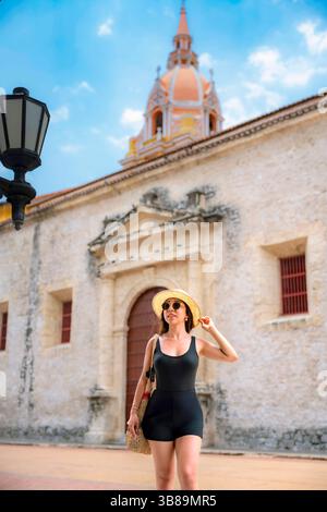 Die junge Latina in schwarzem Strampler, Strohhut und Sonnenbrille steht freudig in der Nähe der berühmten Kirche in Cartagena, Kolumbien. Stockfoto