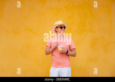 Ein stilvoller junger Latino-Mann mit Korallenhemd und Strohhut lächelt, während er vor der leuchtend gelben Wand in Cartagena, Kolumbien, Saft hält. Stockfoto