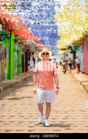Ein junger, lächelnder Latino-Mann geht auf einer geschmückten Straße in Getsemaní, Cartagena, mit Korallenhemd, weißen Shorts, Strohhut und Sonnenbrille Stockfoto